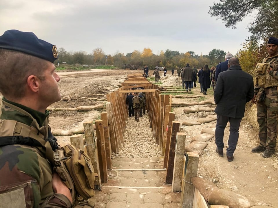 Soldaten neben einem Schützengraben, Menschen gehen vorbei.