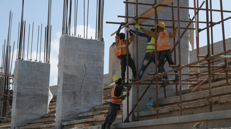 Bauarbeiter auf einer Baustelle mit Gerüsten, Betonpfeilern und Stahlträgern.