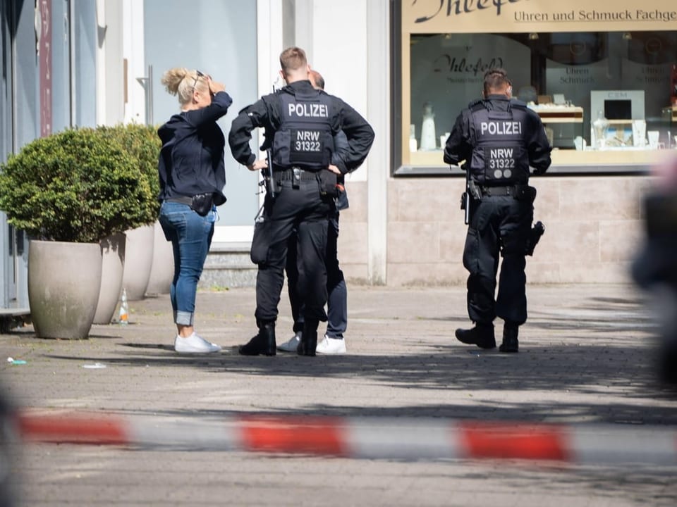 Mehrere Polizisten stehen auf einer Strasse, reden mit einer Person, Absperrband im Vordergrund.