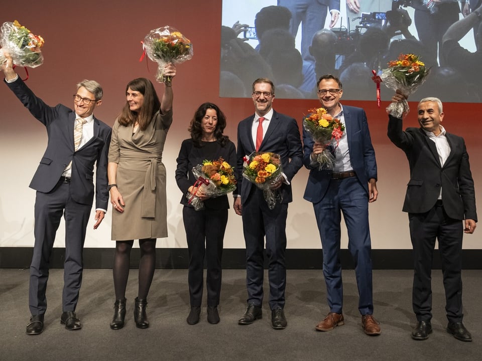 Sechs Leute winken auf Bühne mit Blumensträussen.