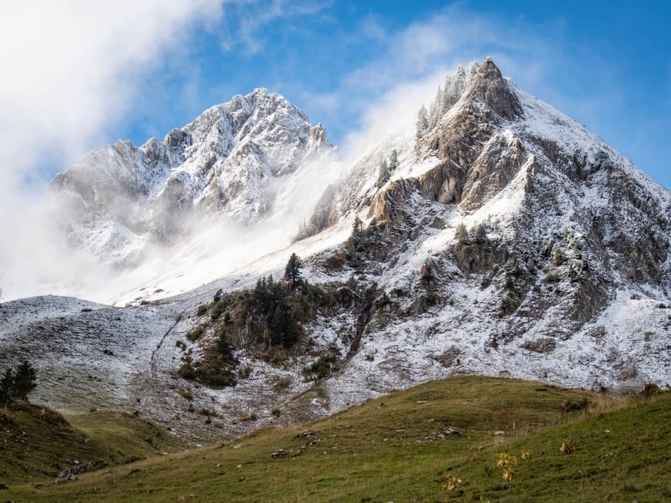 Berggipfel mit Schnee und Wolken unter blauem Himmel.