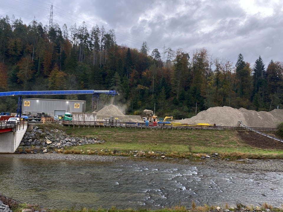 Baustelle am Flussufer mit Bagger und Kieshaufen.