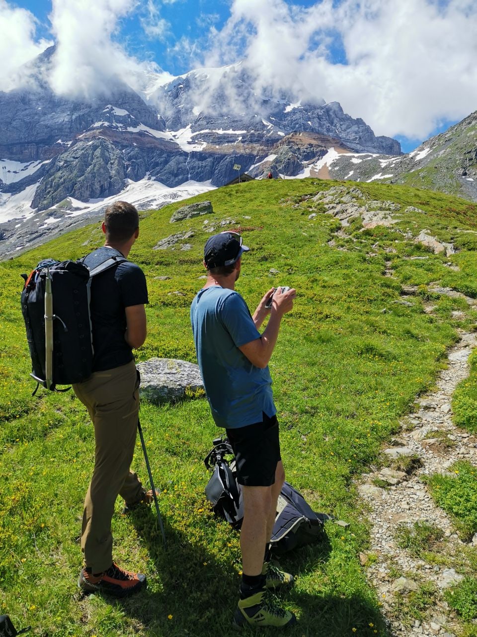 Zwei Wanderer auf Gebirgspfad mit verschneiten Bergen im Hintergrund.