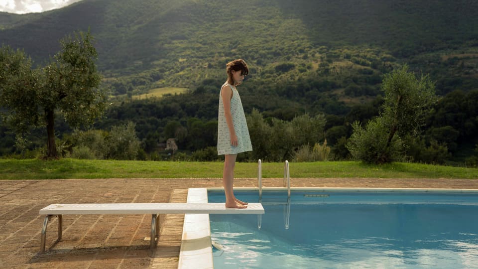 Eine junge Frau steht in einem Sommerkleid auf einem Pool-Sprungbrett vor einer hügelig-ländlichen Kulisse.