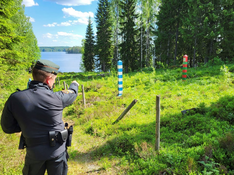 Ein Grenzbeamter zeigt auf Markierungen im Wald am Seeufer.