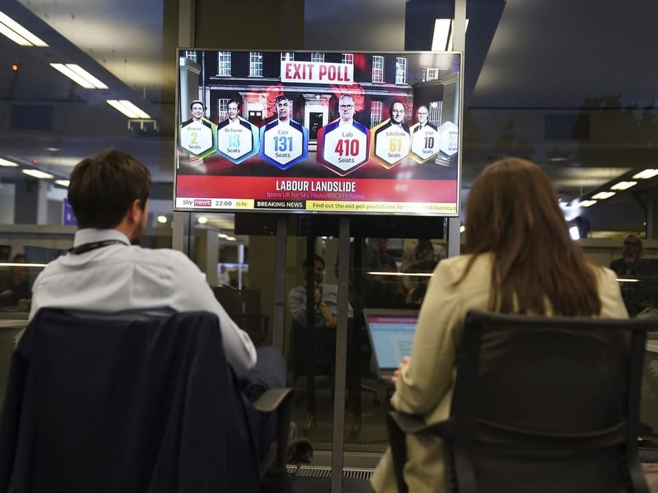 Zwei Personen sehen eine Wahlberichterstattung auf einem Monitor.