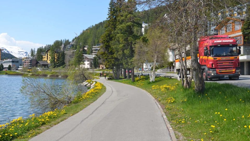 Asphaltierter Weg entlang des St. Moritzer Sees, mit Blumen und Bäumen, und einem roten Lastwagen auf der Strasse.