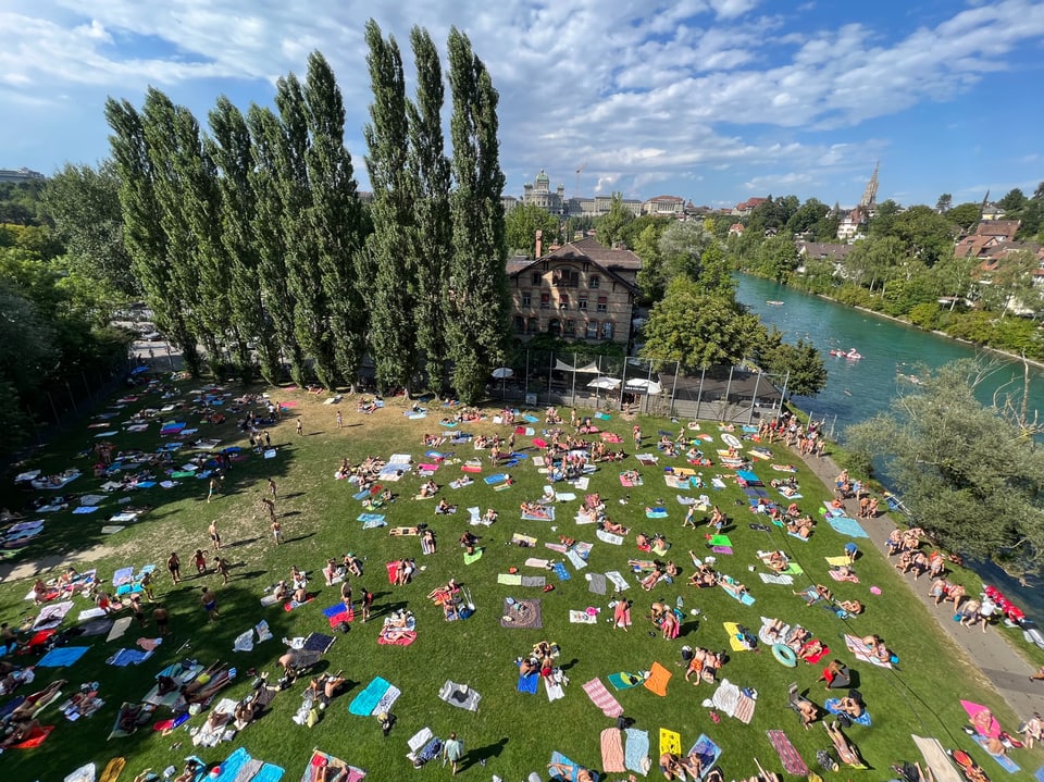 Luftaufnahme eines Parks mit vielen Menschen auf Liegestühlen und Decken neben einem Fluss.