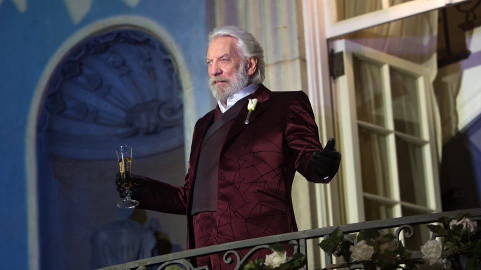 Donald Sutherland steht im Samtanzug auf einem Balkon und hält ein Glas Sekt in der rechten Hand.