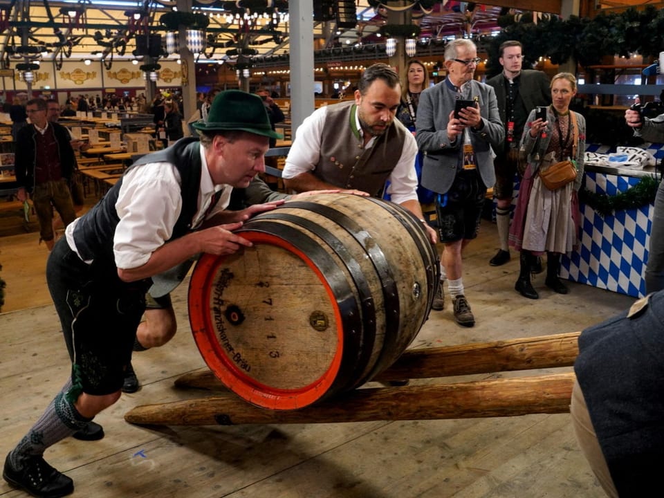 Zwei Männer rollen ein Bierfass auf einem Holzgestell bei einem Fest.