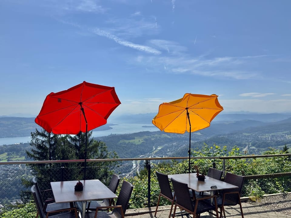 Terrasse inklusive gelben und roten Sonnenschirm mit Blick Richtung Zürichsee bei blauem Himmel.
