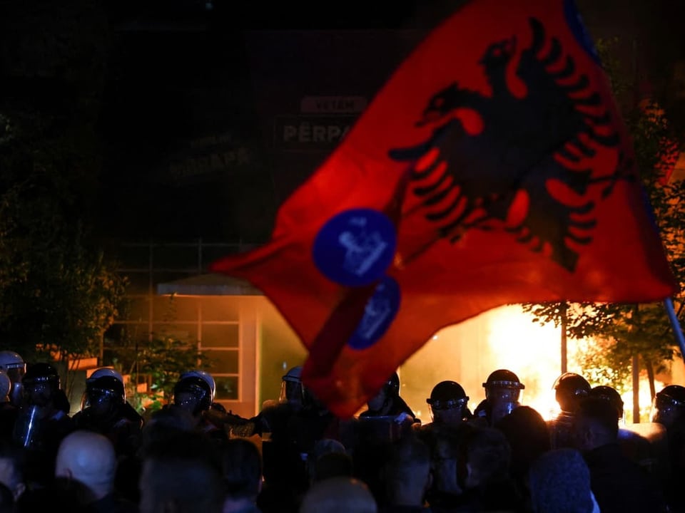 Demonstration mit albanischer Flagge bei Nacht, Feuer im Hintergrund.