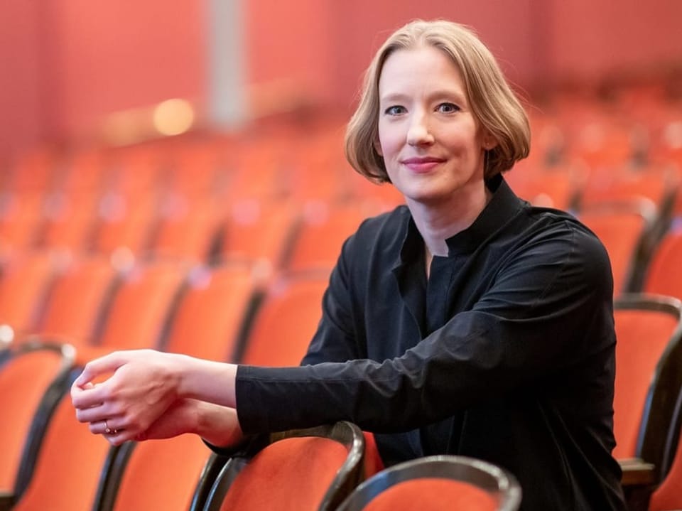 Frau mit kurzen blonden Haaren sitzt in einem leereren Opernhaus mit roten Sitzen.