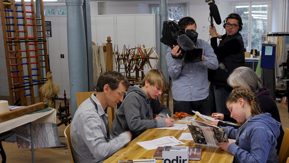 Familie mit zwei Kindern sitzt am Tisch, füllt Fragebogen aus. Kamerateam im Hintergrund.