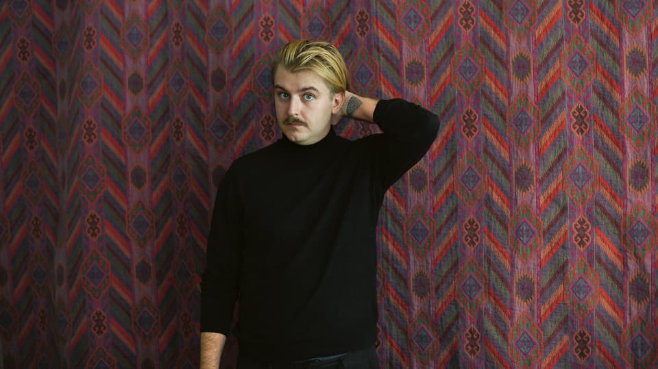 Porträtbild von Artur Weigandt mit schwarzem Pullover vor rötlich gemustertem Hintergrund