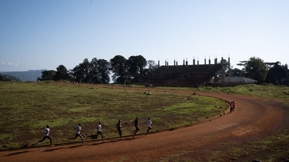 Sechs  Läuferinnen und Läufer auf dem Kamariny Track. Einige Säulen der unvollendeten Tribüne ragen in den Himmel.