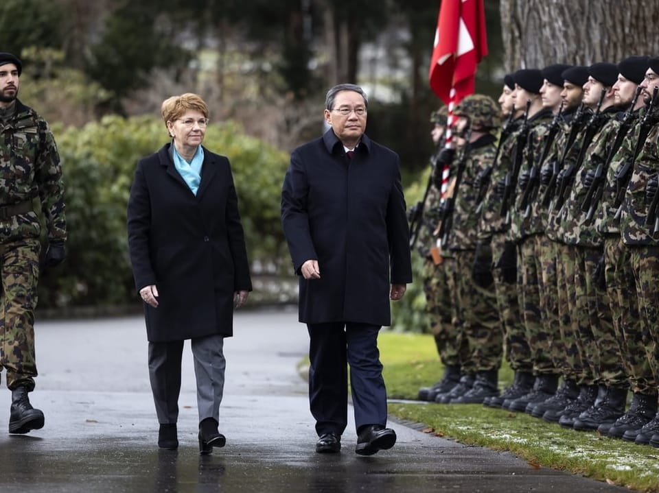links eine Frau im Mantel, rechts ein Mann mit Brille. Laufen draussen an Militärreihe vorbei