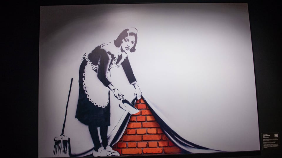Ausstellungsuafnahme eines Graffiti von Banksy. Ein Dienstmädchen hält eine Schippe, die andere Hand hebt die Tapete.