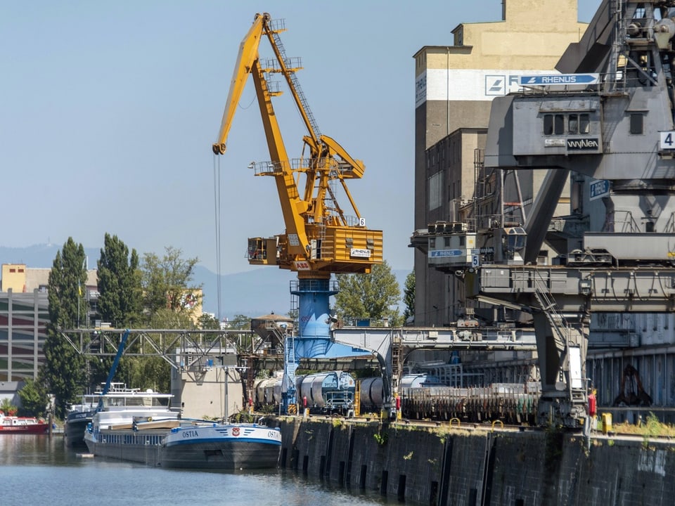 Im Vordergrund treibt ein Frachtschiff im Rhein, dahinter stehen Güterzüge auf Schienen und ein grosser Kran.