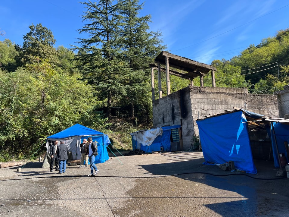 Menschen stehen vor blauen Zelten und einer verfallenen Betonstruktur im Freien.