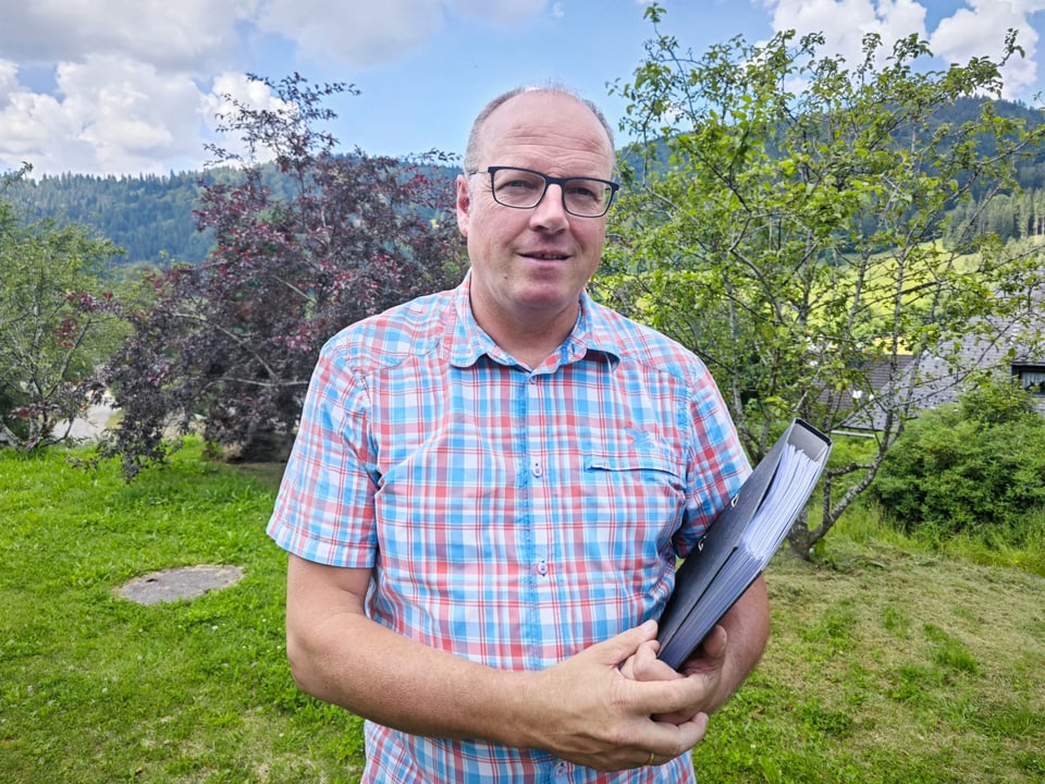 Ein Mann steht in einem Garten. In der Hand hält er einen schwarzen Ordner. 