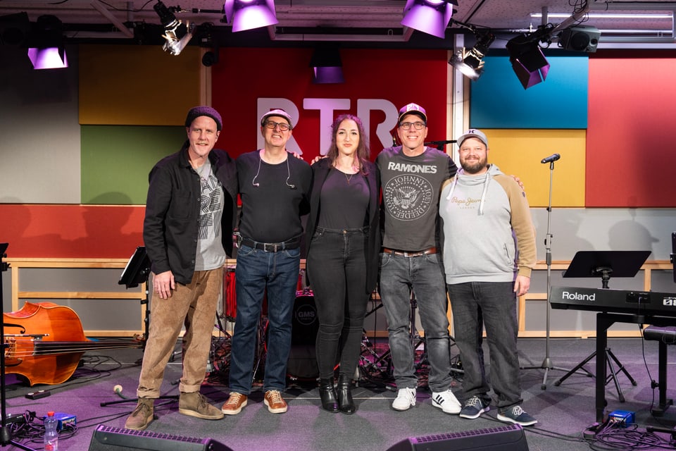 Fünf Personen posieren in einem Musikstudio vor einer farbigen Wand.