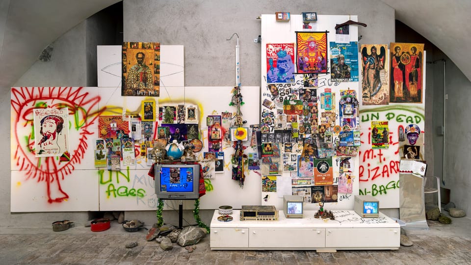 Ausstellungsansicht von Lee «Scratch» Perrys Werk: viele kleinteilige Objekte hängen an einer Wand, dazu Farbsprüherei.