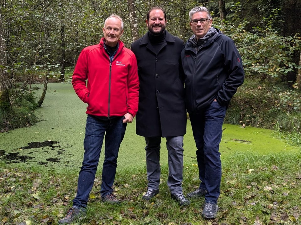 Drei Männer stehen im Wald vor einem Teich. 