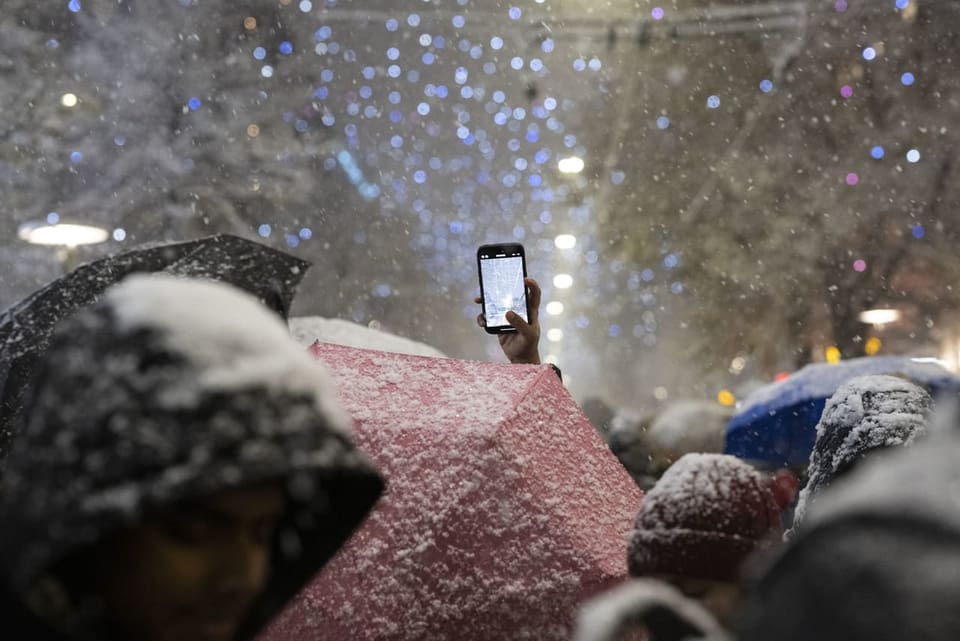 Menschen mit Schirmen im Schneefall, jemand hält ein Handy hoch.