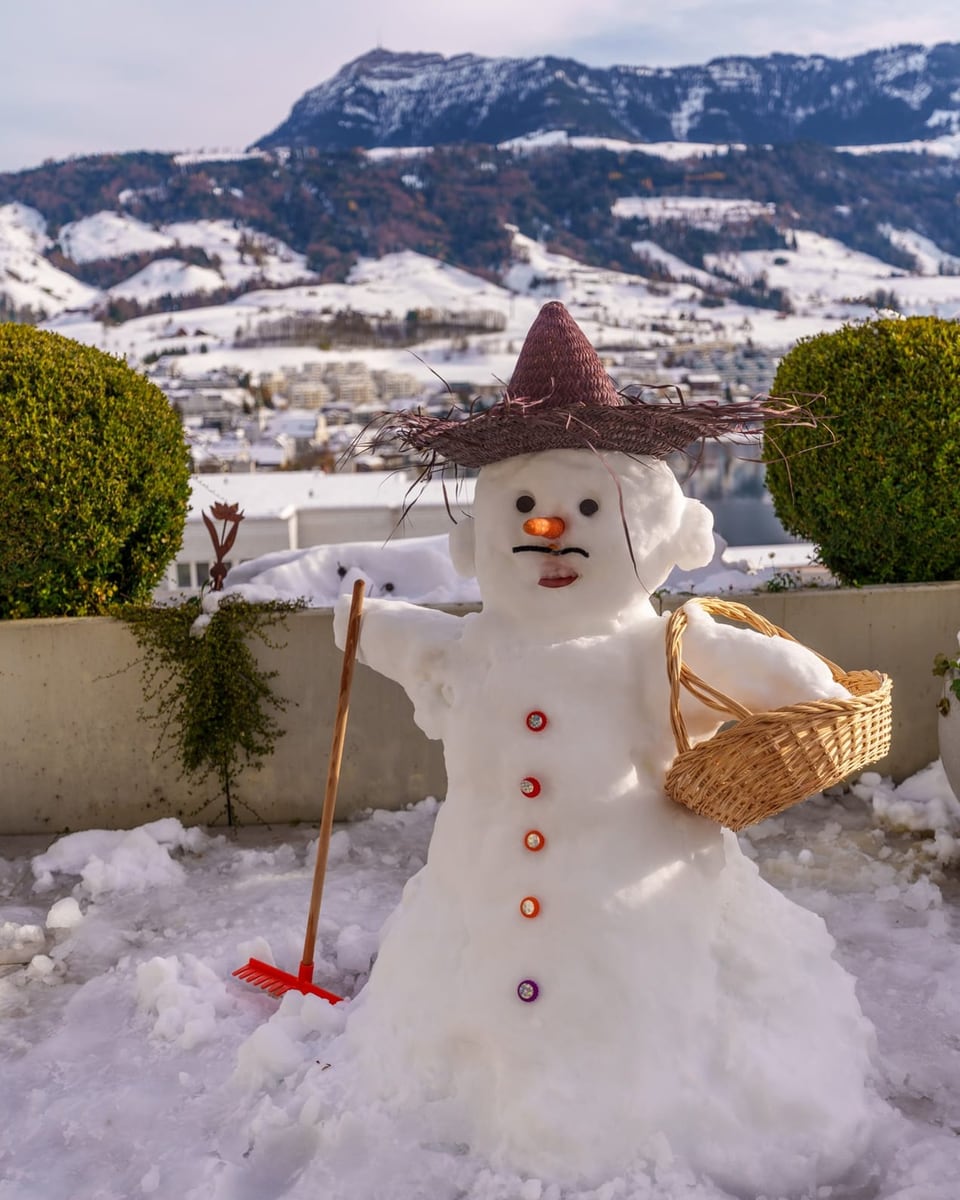 Schneemann mit Hut und Korb vor schneebedeckten Bergen.