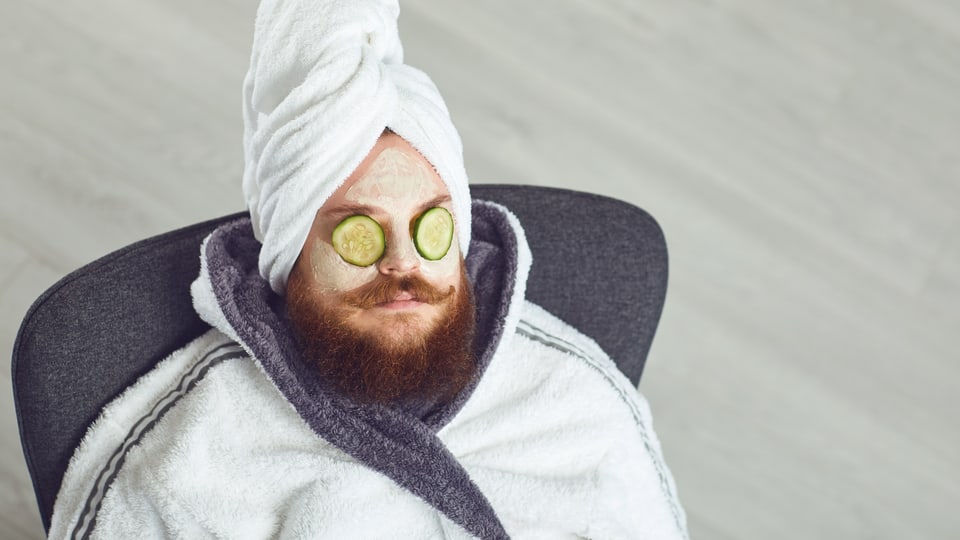 Ein Mann in Bademantel, mit Bart, zwei Gurkenscheiben auf den Augen und Gesichtscreme.