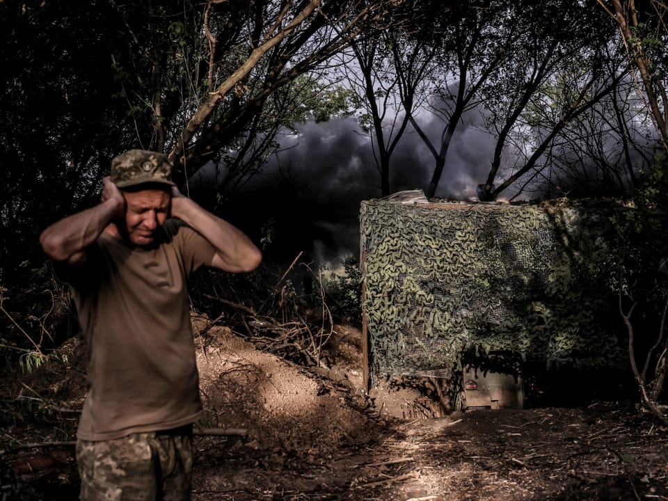 Soldat hält sich die Ohren zu, Explosion im Hintergrund.