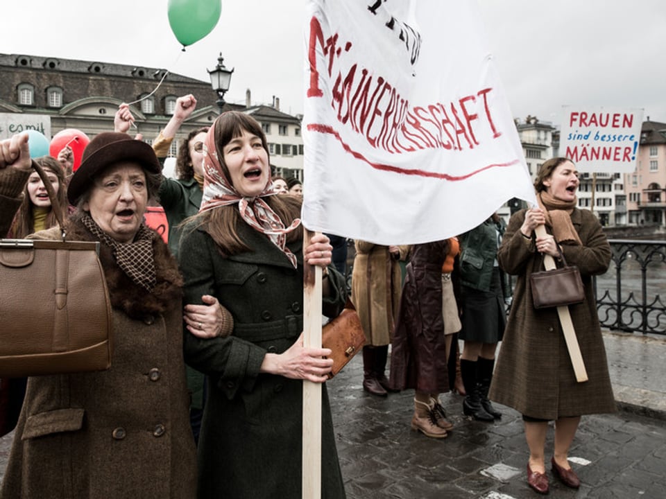 Filmstill: Frauen demonstrieren auf einer Brücke das Frauenstimmrecht.