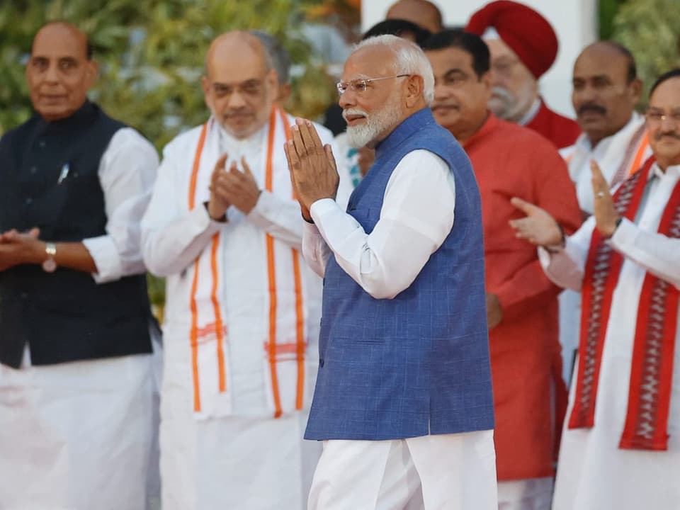 Modi in blau-weissem Gewand, Hände vor der Brust, läuft an Menschenmenge vorbei (verschwommen im Hintergrund).