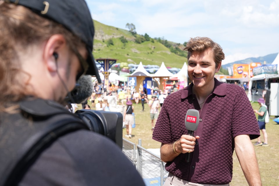 Reporter mit Mikrofon im Freien, Kameramann im Vordergrund.