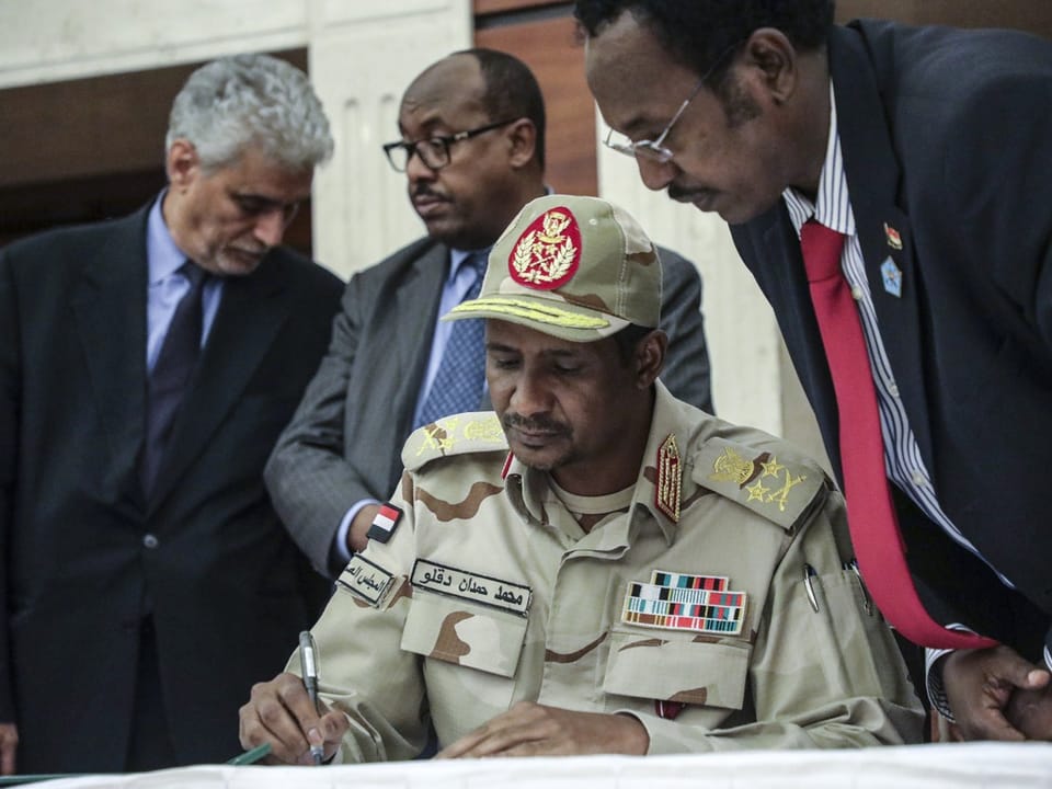 Mann in Militäruniform, der ein Dokument unterschreibt, umgeben von drei Männern.