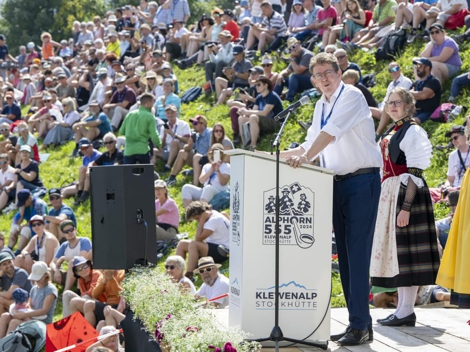 Rösti at the lectern. Behind him, many guests are sitting on the lawn and seem to be listening.