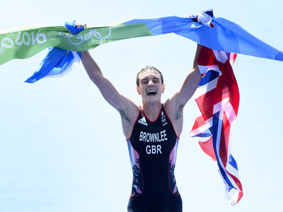 Triathlet hebt Union Jack und Olympische Flagge hoch, strahlend vor Freude.