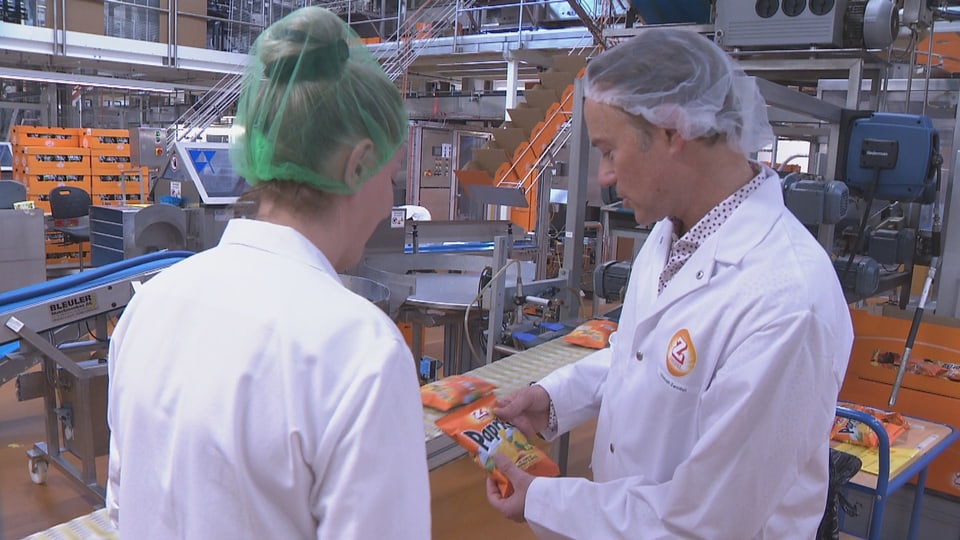 Zwei Personen in Laborkitteln und Haarnetzen in einer Fabrik besprechen Chips-Verpackung.