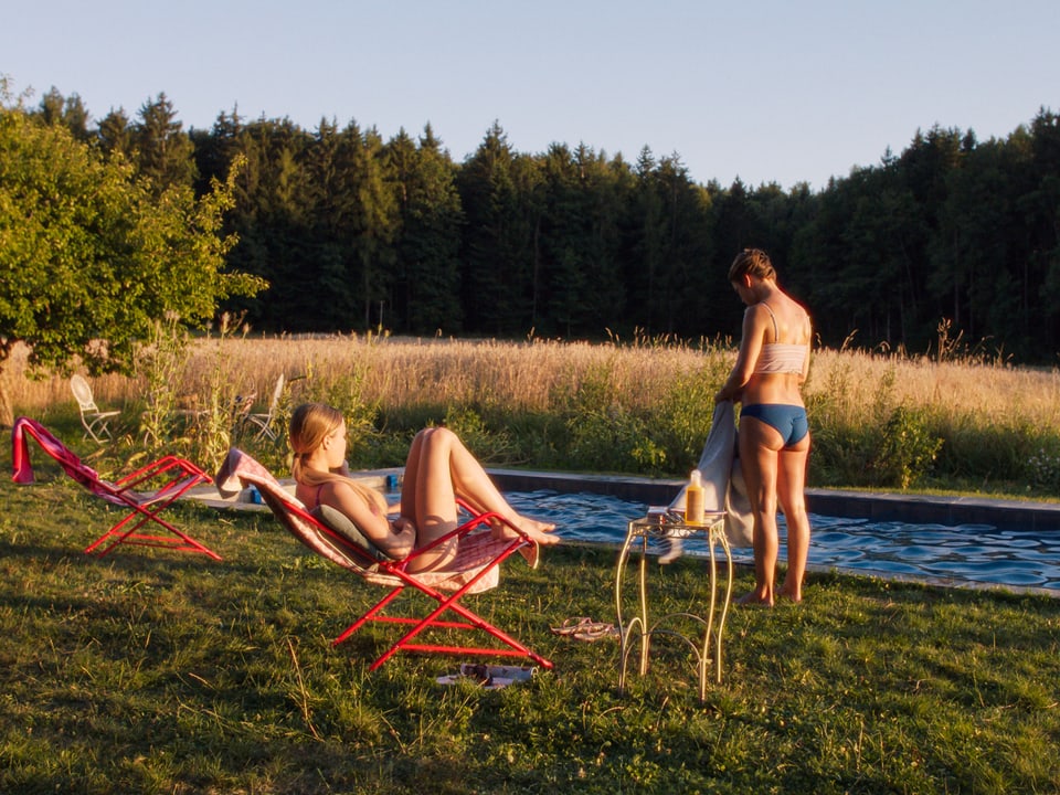 Swimmingpool, Liegestühle, eine Person sitzt, eine andere steht, goldenes Licht.