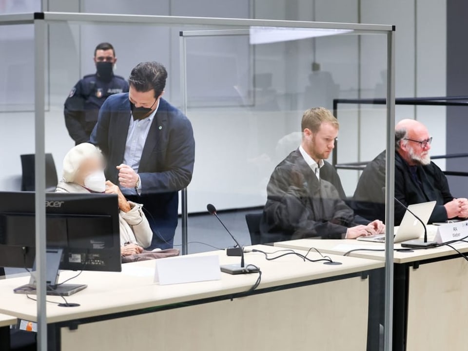 Hinter einer Glasscheibe sitzt die Angeklagte neben ihren zwei Anwälten.