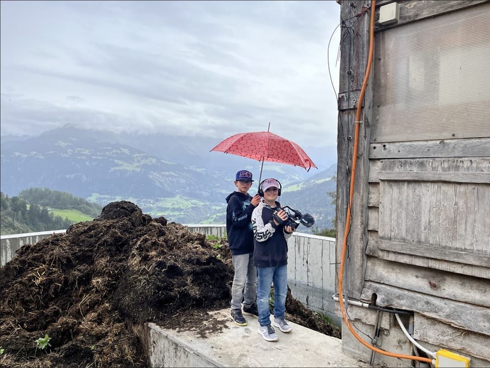 Zwei Kinder mit rotem Regenschirm stehen neben einem Holzhaufen vor einem Bergpanorama.