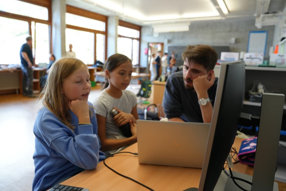 Erwachsener unterrichtet zwei Kinder am Computer in einem Klassenzimmer.