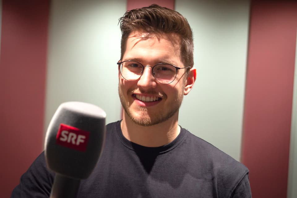 Mann mit Brille hält SRF-Mikrofon vor gestreiftem Hintergrund.