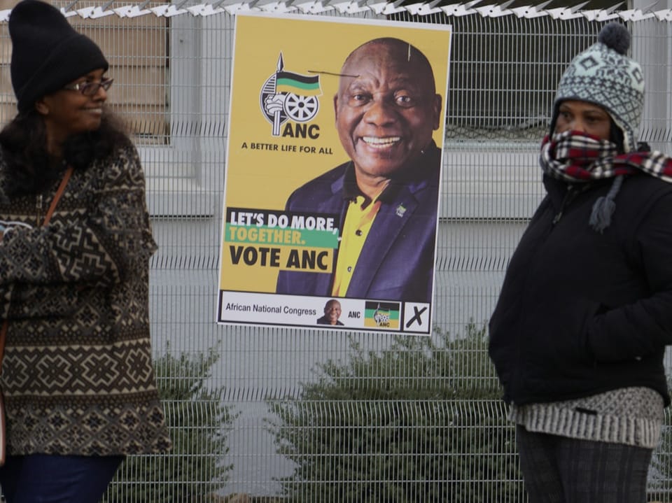 Zwei Frauen stehen in dicke Kleidung gehüllt neben einem Wahlplakat von Cyrill Ramaphosa