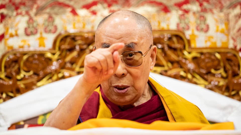 Der Dalai Lama deutet mit dem Zeigefinger in die Luft.