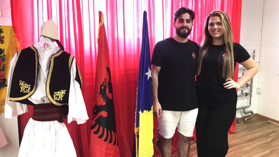 Ein Mann und eine Frau stehen neben einer Flagge von Albanien und einem Anzug aus weissem Stoff. 