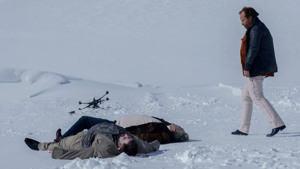 Zwei tote Männer liegen im Schnee neben einer Drohne, ein Mann in Lederjacke läuft auf sie zu
