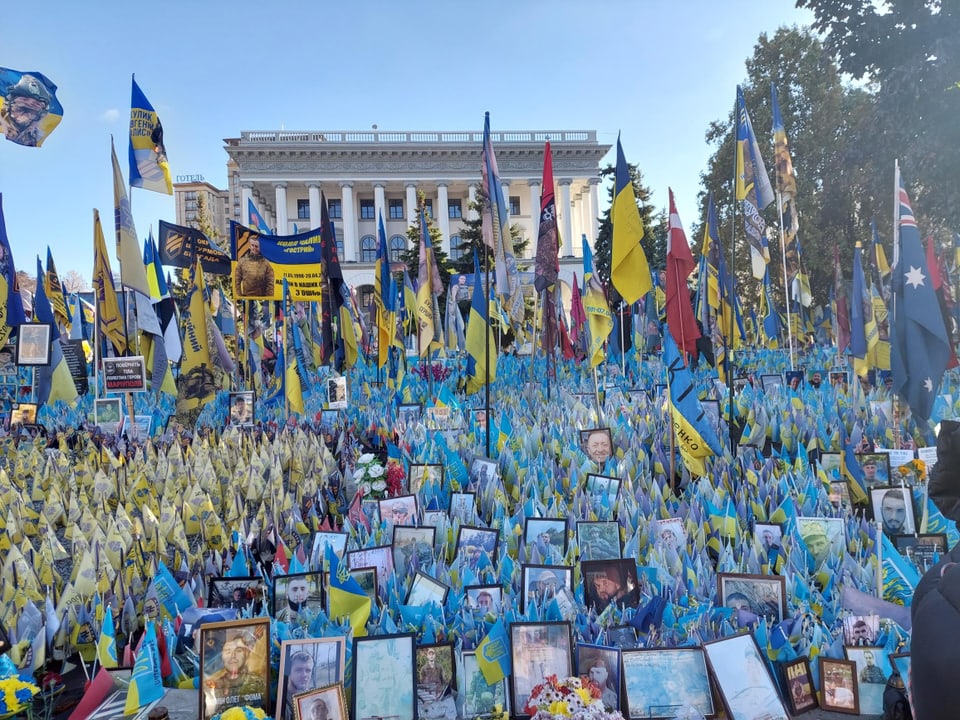 Hunderte blau-gelbe Flaggen und Fotos, Gebäude im Hintergrund.