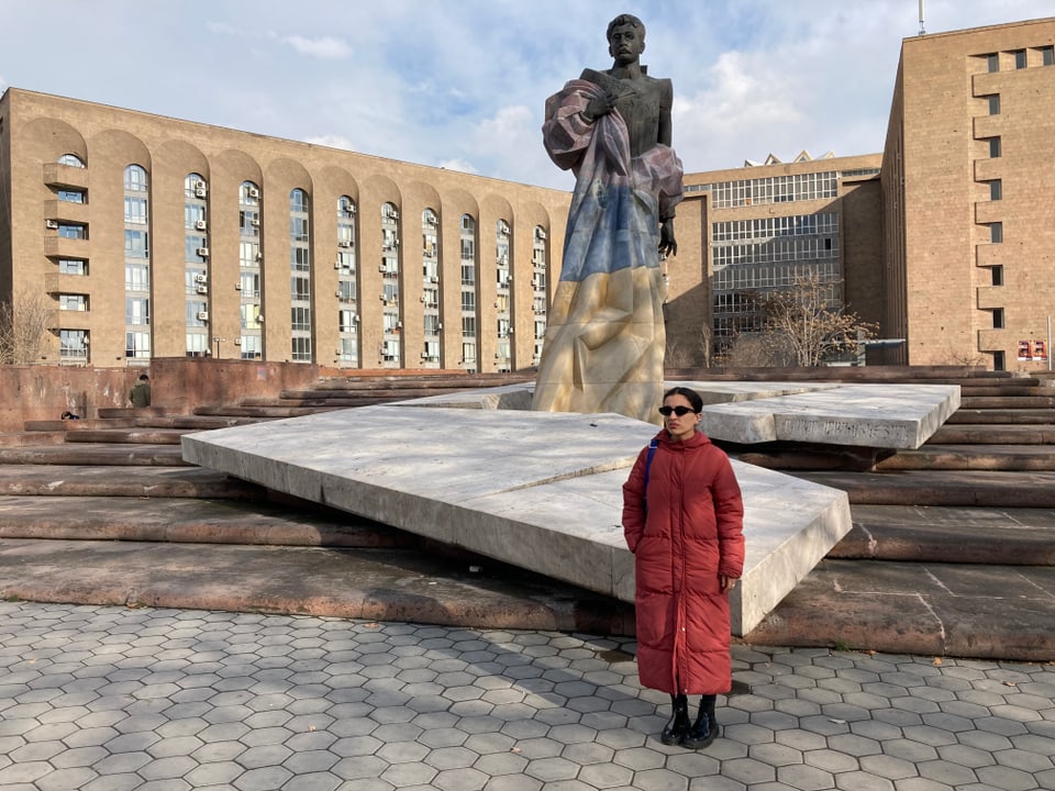 Eine Frau in rotem Daunenmantel steht neben einer Statue
